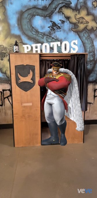 The image features a man dressed as a character from the video game Minecraft, standing in front of a wooden photo booth with a sign that says "PHOTOS" above him. The man appears to be posing for a picture or participating in a photoshoot within this themed setting.