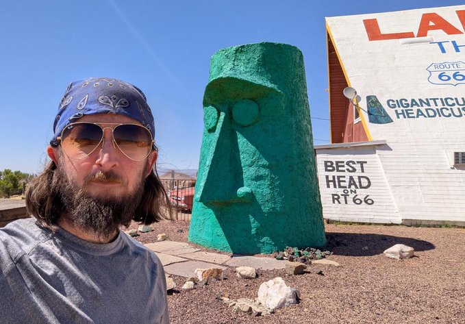 The image features a man wearing sunglasses and a bandana around his neck, standing next to a green statue of an old man's head with a beard. The statue appears to be made out of cement or stone and is located outdoors. In the background, there are two buildings - one being a house and another a barn. A sign can also be seen in the image that reads "Cardano".