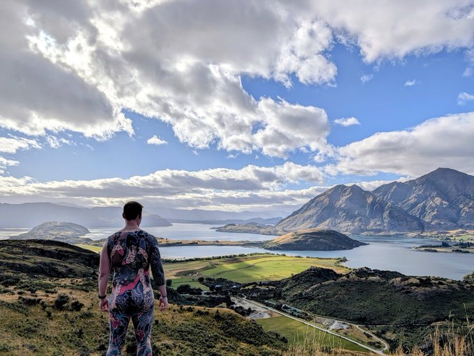 A man with a patterned backpack overlooks a scenic mountain landscape with a lake and green fields under a cloudy sky.