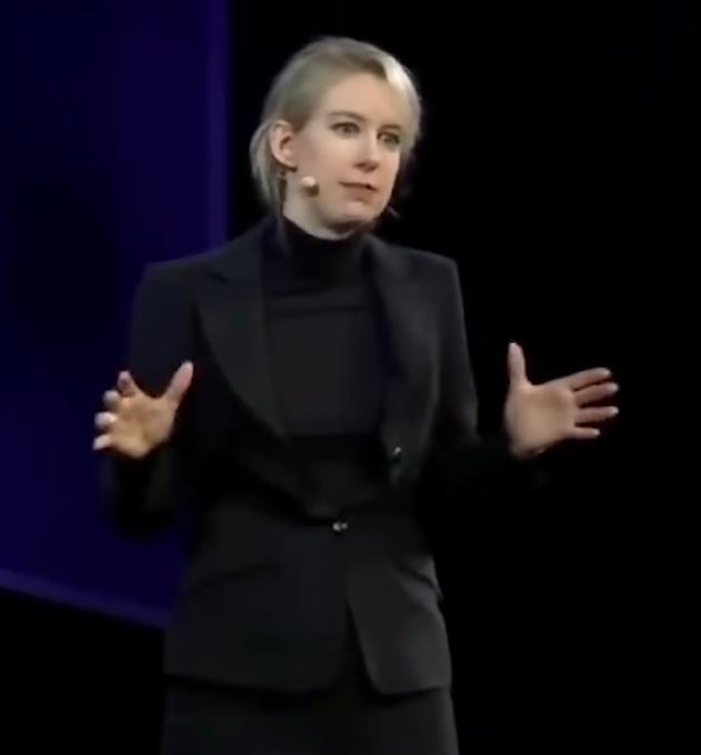 Elizabeth Holmes, dressed in a black blazer and turtleneck with a headset microphone, gestures expressively with both hands while speaking on stage—her familiar public speaking style, here repurposed to humorously echo the post's suggestion to rebrand NFTs with a more trustworthy image.