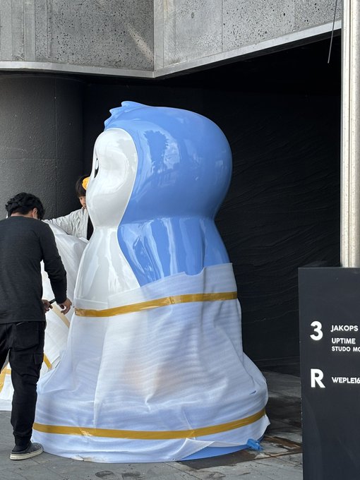 Two workers are installing or handling a large inflatable penguin mascot outside a building marked '3 JAKOPS UPTIME STUDIO MON' and 'WEPLEMEN'.