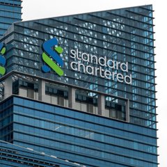 A photograph of a tall modern office building featuring the 'Standard Chartered' logo with its distinctive blue and green intertwined symbol on the glass facade.