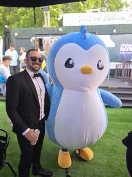 A man in a black tuxedo and bow tie poses for a photo next to an inflatable penguin mascot at an outdoor event.