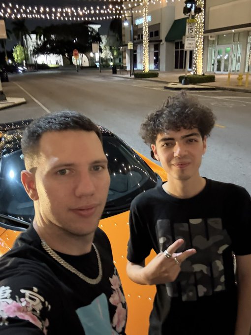 Two young men pose for a nighttime selfie in front of a bright orange McLaren supercar on a city street decorated with string lights. The driver holds up a peace sign while showcasing his new "whip," fitting the celebratory post congratulating @pr6spr on the flashy memecoin-funded purchase.