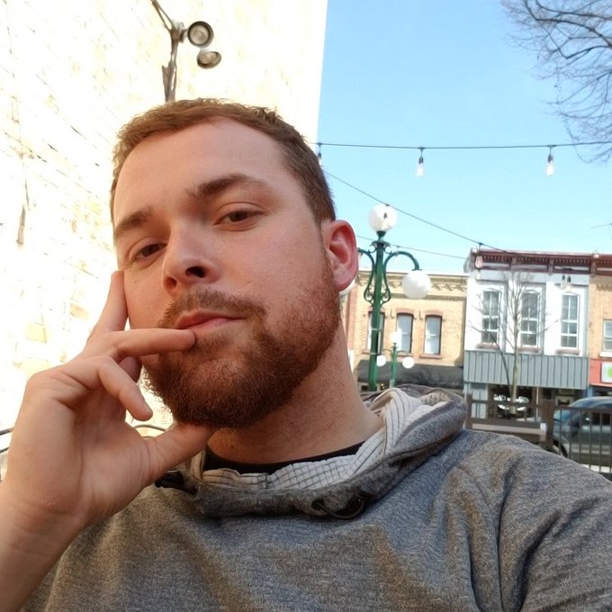 A bearded man with a thoughtful expression rests his hand on his chin in a selfie taken outdoors, with string lights and brick buildings visible in the background.