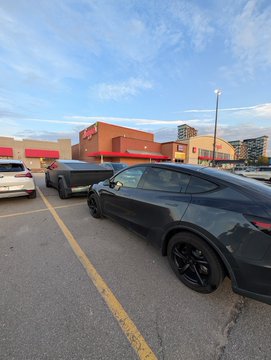 A dark grey Tesla Model Y is parked in a parking lot outside what appears to be a Costco warehouse store.