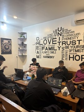 A group of people sit around a table with laptops in a room decorated with a large word-art wall mural featuring motivational words like 'LOVE,' 'FAMILY,' 'JUST,' and 'HOPE.'