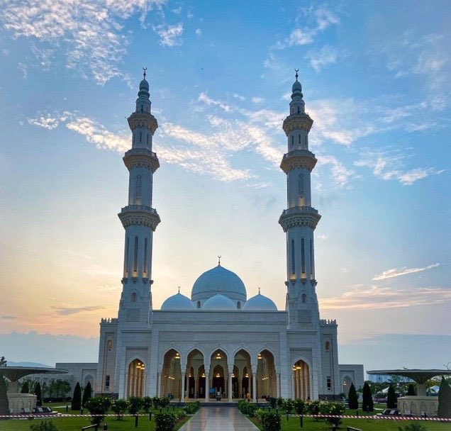A large white mosque with two tall minarets and several domes stands at sunset under a blue, partly cloudy sky.