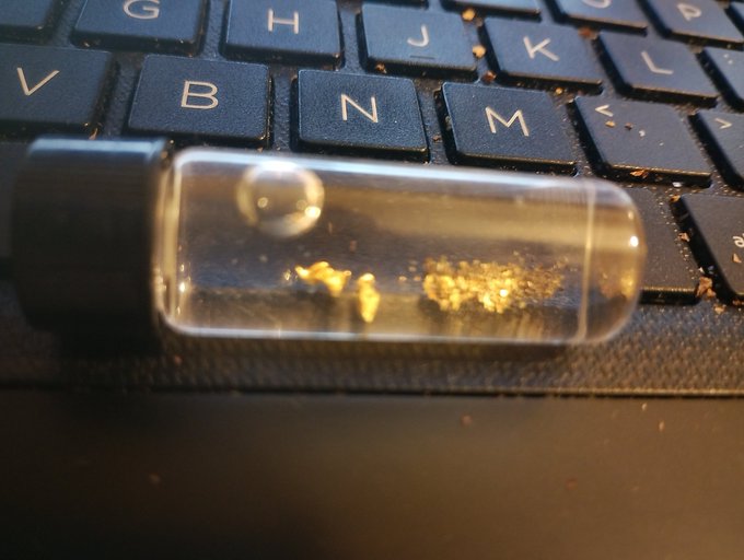 A small clear glass vial with a black cap containing tiny gold flakes rests on a black laptop keyboard.