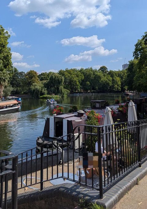 A scenic view of a lake with boats including swan-shaped paddle boats, willow trees, and an outdoor dining area with umbrellas and flower planters enclosed by a black metal fence.
