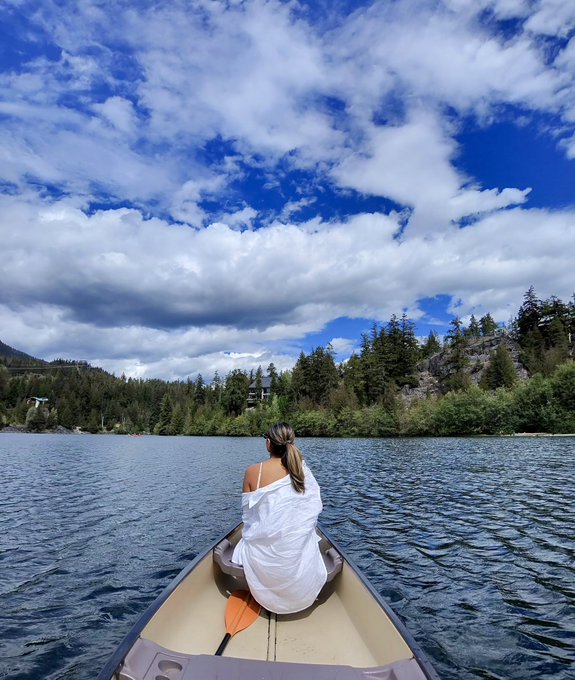 A woman in a white cover-up sits in the bow of a canoe holding an orange paddle, looking out across a lake toward a forested shoreline under a cloudy blue sky.
