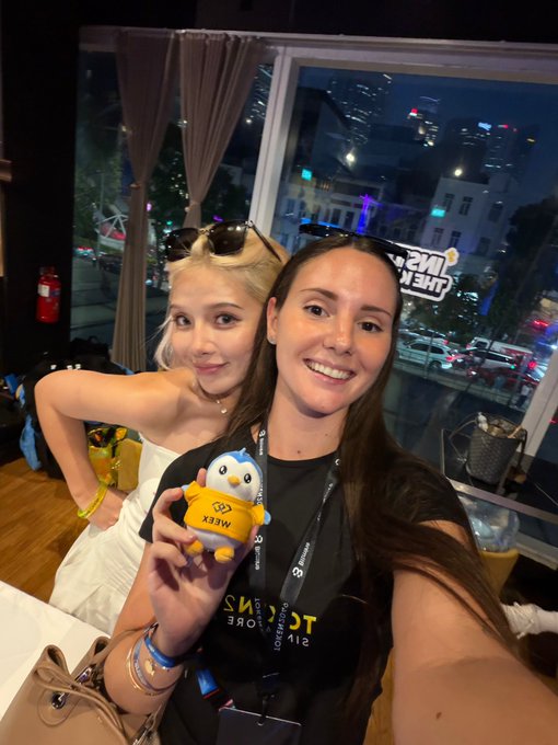 Two women take a selfie indoors with one holding a yellow penguin plush toy labeled 'MEGA', with a window showing a city street at dusk in the background.