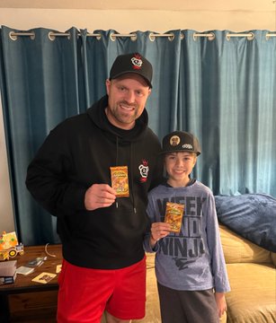 A man wearing a black hoodie and red shorts stands next to a smiling boy in a purple shirt and baseball cap, both proudly holding Pokemon trading cards in a living room.