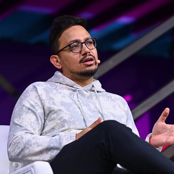 The image shows a man wearing glasses sitting down with his hands outstretched, possibly gesturing or making a point during a conversation. The background features purple and blue colors, which could be part of an event setting or stage design. The caption provided in the image suggests that the main focus is on the man's actions and gestures rather than the surrounding environment.