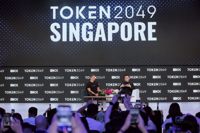 Two men sit on stage at a 'TOKEN 2049 SINGAPORE' conference, with the event name and 'OKX' logos displayed on large screens behind them.
