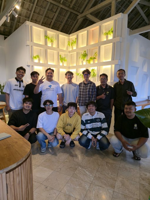 A group photo of eleven men posing together indoors in front of a white wall with recessed shelves displaying plants.
