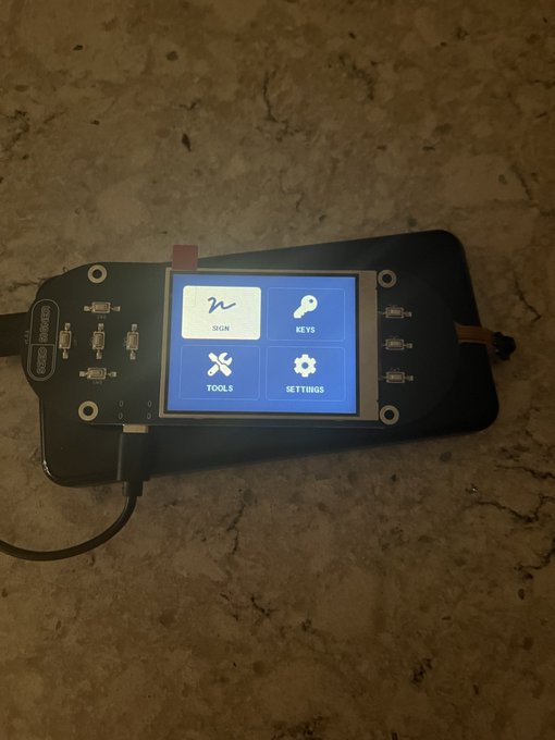 A black electronic device with multiple ports, labeled "SPACEHACKER", lies on a stone surface, displaying a screen with four options: "SIGN", "KEYS", "TOOLS", and "SETTINGS".