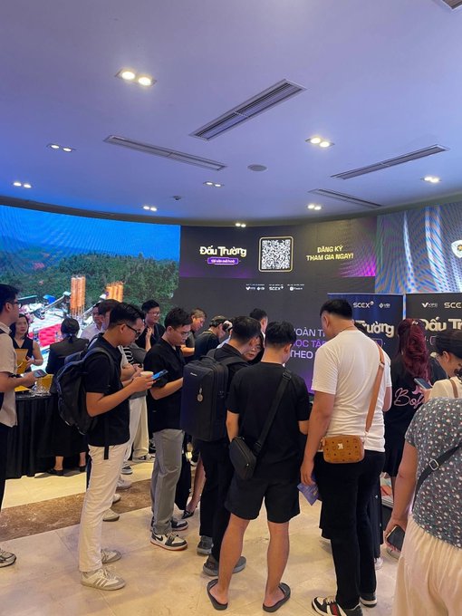 Photo of a crowd at an indoor event with a Vietnamese banner reading 'Dấu Trường', a QR code, and 'DANH SÁCH ĐẦU TƯ' (Investor List).