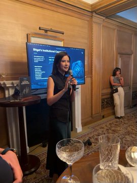 A woman in a dark sleeveless top presents at an institutional event, standing beside a slide titled "Bitget's Institutional C..." while another attendee looks on from the background, visually capturing the speaker's address to the 60+ institutional partners mentioned in the post.
