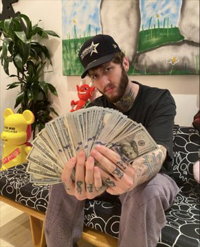 A heavily tattooed man wearing a black Dallas Cowboys cap fans out a handful of U.S. dollar bills, sitting in a room decorated with yellow Homer Simpson figures, a Lucky Charms cereal box, and cow-patterned cushions. The image appears to illustrate the "massive backlash" FaZe Banks received after promoting a memecoin that reportedly lost around 85% of its value, contrasting the cash he displays with the losses suffered by followers.