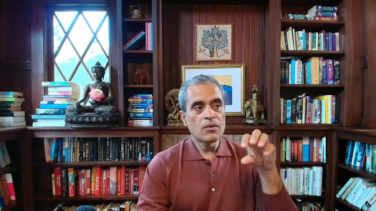 The image shows an older man sitting at a desk surrounded by numerous books. The man appears to be engaged in a video call or presentation, as he points towards something off-camera. The background features a wooden bookshelf filled with various books of different sizes and colors. The presence of the bookshelf suggests that this scene takes place in an environment where learning, reading, or research is common.