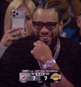 The image features a man wearing glasses, smiling at the camera while holding up his cell phone. He appears to be taking a picture of himself or someone else with the phone's front camera. The background shows that he is surrounded by other people who are also present in the scene. There is an element related to sports in the image as well, which could indicate that this gathering might be at a sporting event where the man and others are fans.

The post on social media seems to be about a basketball game or match between two teams, with one team losing some money. The caption reads "Solkrow lost some good money today". This suggests that the image captures an interesting moment during a sports event, possibly involving the man in glasses and his friends who are also present at the gathering.
