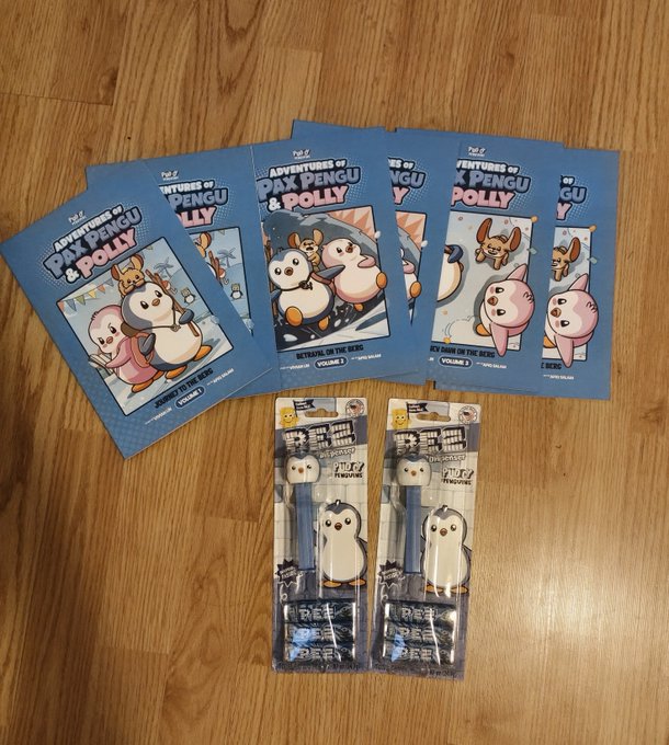 Five blue folders titled 'ADVENTURES OF PAX PENGU & POLLY' featuring cartoon penguins and a pink cat are scattered on a wooden floor, alongside two packaged pens with matching penguin designs.