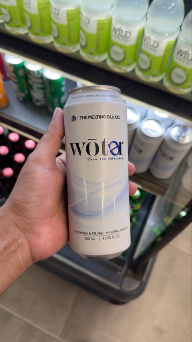 A hand holds up a can of "WŌTA Premium Natural Mineral Water" from "The Melting Blu Co." in a 500 mL (16.90 fl oz) size, labeled as "A Joyful Collaboration in Every Sip," in front of a store shelf stocked with "WILD" branded beverages, perfectly tying into the post's friendly reminder to "GM! Reminder to drink some water."