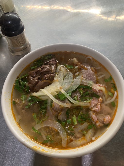 A bowl of Vietnamese-style beef noodle soup (pho) topped with sliced beef, onions, and fresh herbs, served on a metal table with a toothpick holder in the corner.