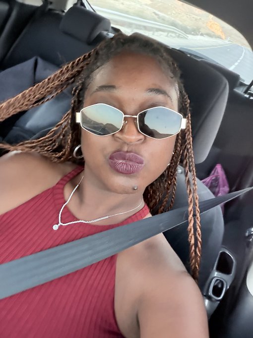 A selfie of a woman with long braided hair wearing geometric sunglasses and a red top, sitting in a car with a seatbelt across her chest.