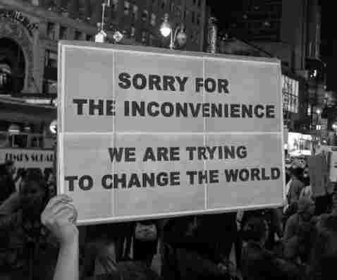 A black-and-white photo of a protest where a person holds a sign reading 'SORRY FOR THE INCONVENIENCE WE ARE TRYING TO CHANGE THE WORLD.'
