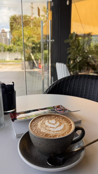 A latte with intricate leaf latte art sits on a saucer at a cafe table by a large window overlooking a city street. The cozy beverage pairs perfectly with a simple "Hello"—a warm invitation to start the day with good coffee and a peaceful view.