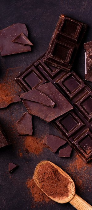 An overhead close-up of dark chocolate bars broken into pieces alongside a wooden spoon filled with cocoa powder, set against a dark, textured surface.