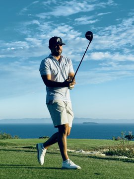 A man wearing a dark cap, sunglasses, grey shirt, and khaki shorts mid-swing with a golf club on a grassy course with the ocean in the background.