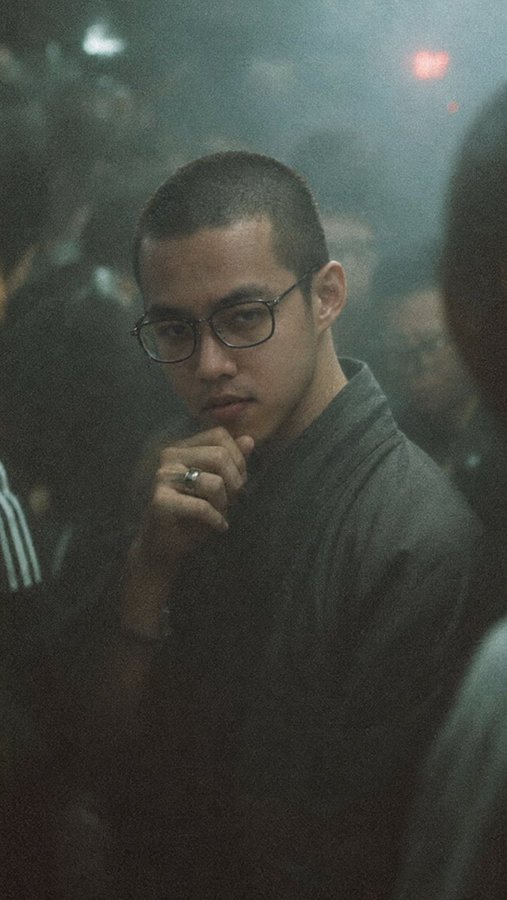 A cinematic portrait of a young man with glasses and short dark hair, wearing a dark shirt, posing thoughtfully with his hand on his chin in a dimly lit setting.
