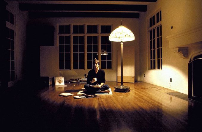 A person sits on the wooden floor of a dimly lit, spacious room illuminated by a single floor lamp, surrounded by scattered papers.