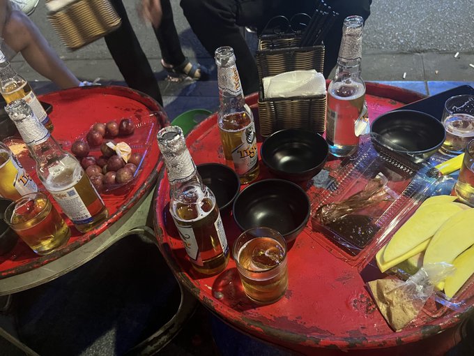 Two red round tables at an outdoor street-side gathering are covered with Asahi beer bottles, small black bowls, glasses of beer, and snacks including grapes and fruit, with people standing around enjoying the drinks. The scene captures the casual weekend hangout described in the post, where friends meet for a few beers before heading home to rest before the work week begins.