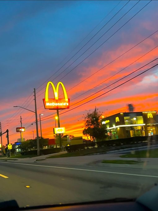 A McDonald's restaurant photographed at sunset, featuring the iconic golden arches sign and a building with the McDonald's logo under a vibrant pink and blue sky.