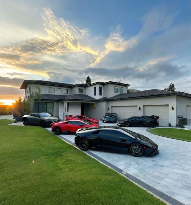 A luxurious modern white mansion at sunset with multiple exotic sports cars including Lamborghinis, Ferraris, and a McLaren parked on the driveway.