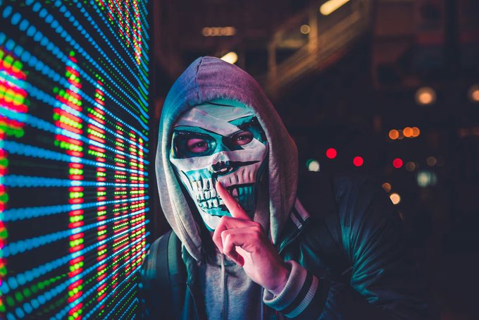 A person wearing a skull mask with red eyes and a gray hoodie holds a finger to their lips in a 'shh' gesture while standing next to a wall of glowing blue, red, and green LED lights.