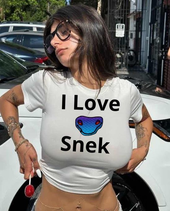 A woman wearing sunglasses and a white "I Love Snek" t-shirt with a blue snake graphic poses in front of a white car, holding a red lollipop while referencing the $SNEK cryptocurrency. The playful snake emoji on the shirt ties directly into the nostalgic $SNEK throwback theme of the post.