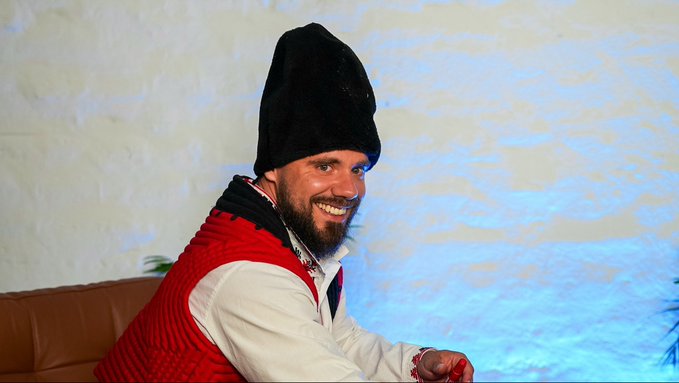 A smiling bearded man wearing a tall black fur hat and a red knitted vest over a white shirt, seated near a blue-lit wall.