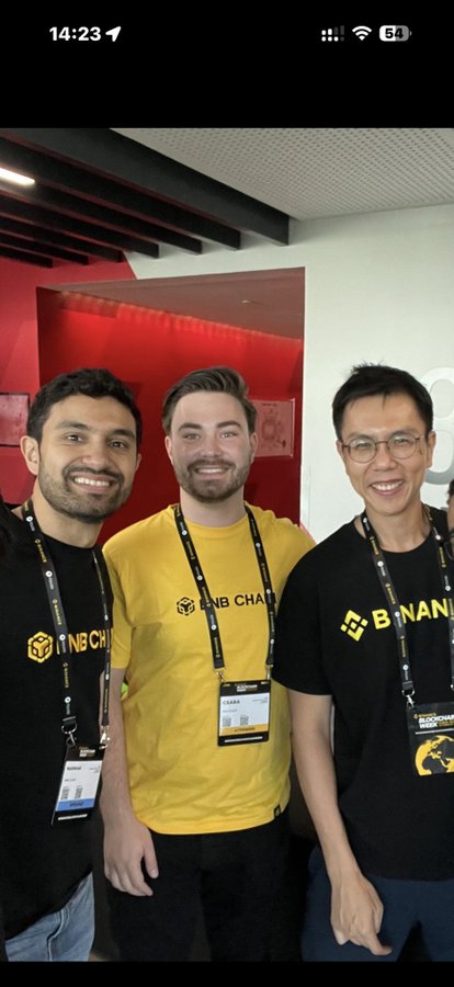 Three men pose together at what appears to be a Binance event, with the center man in a yellow "BNB Chain" shirt flanked by two others in black Binance-branded shirts, all wearing lanyards and badges. The image relates to the post by showing apparent Binance representatives, contextualizing the poster's commentary about CZ and the crypto community's stance on a scam.