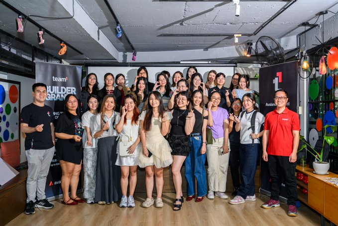 A group photo of team members posing together at what appears to be a T1 (team builder program / Avalanche event) gathering, with banners visible in the background.
