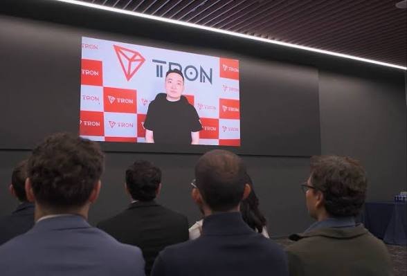An audience viewing a large screen displaying the TRON cryptocurrency logo with a person in a black shirt speaking, while attendees in business attire watch from their seats in what appears to be a corporate event.