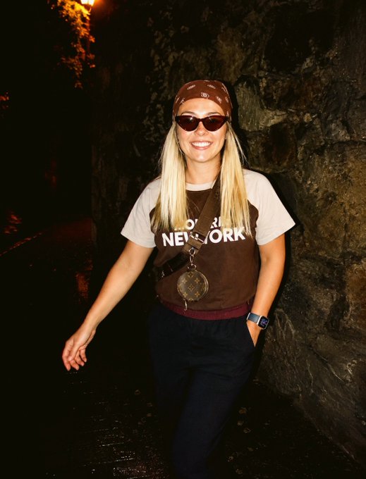 A blonde woman wearing sunglasses, a brown bandana, and a 'NEW YORK' t-shirt smiles while standing near a rock wall.