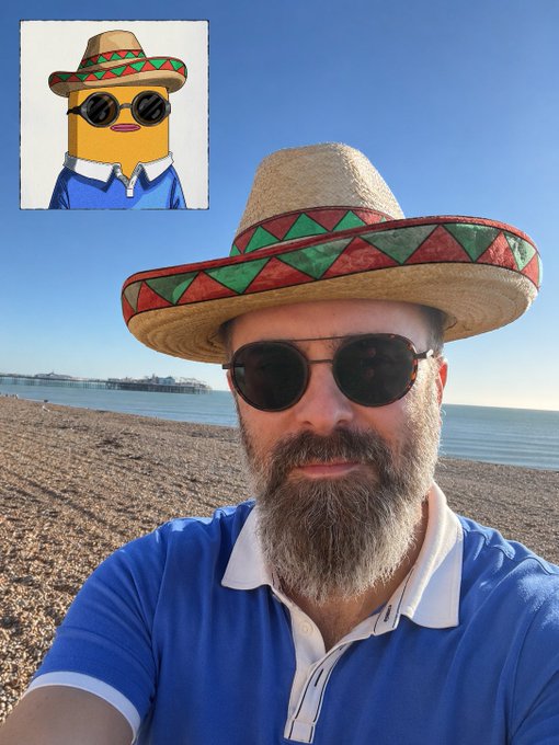 A beach selfie of a bearded man wearing a blue polo shirt, dark sunglasses, and a straw sombrero with a red and green patterned band; a small inset image in the upper left shows a cartoon duck avatar dressed identically.