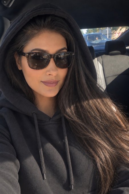 A woman with long dark hair wearing a black hoodie and dark sunglasses takes a selfie while sitting in a car.