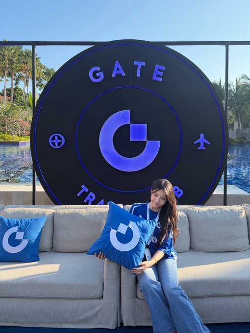 A woman wearing a blue t-shirt and jeans sitting on a beige couch holding a blue 'G' logo pillow, with a large circular 'GATE TRADE' sign behind her.