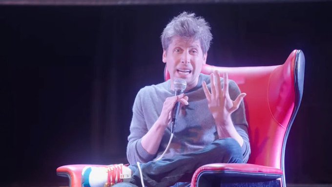 Sam Altman sits in a red leather chair holding a microphone, appearing animated and expressive during what looks like a panel discussion or interview, his visibly stressed demeanor aligning with the post's joke about his recent aging under pressure at OpenAI.